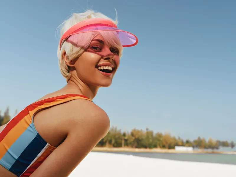 Portrait of a smiling woman on a sunny beach, wearing a colorful striped swimsuit and pink visor, with a bright blue sky and calm water in the background.