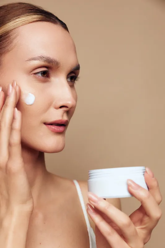 Closeup of a smiling woman applying moisturizer to her cheek, holding a jar. Soft neutral backdrop emphasizes skincare, self care, and a calm beauty routine with warm lighting