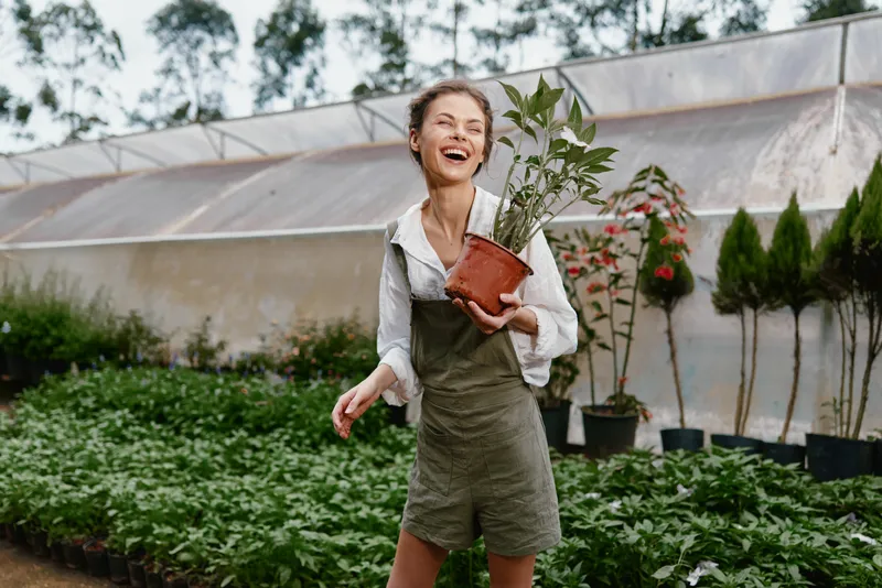 Happy young woman in gardening overalls holding a potted plant in a greenhouse filled with greenery, representing joy and connection to nature during gardening