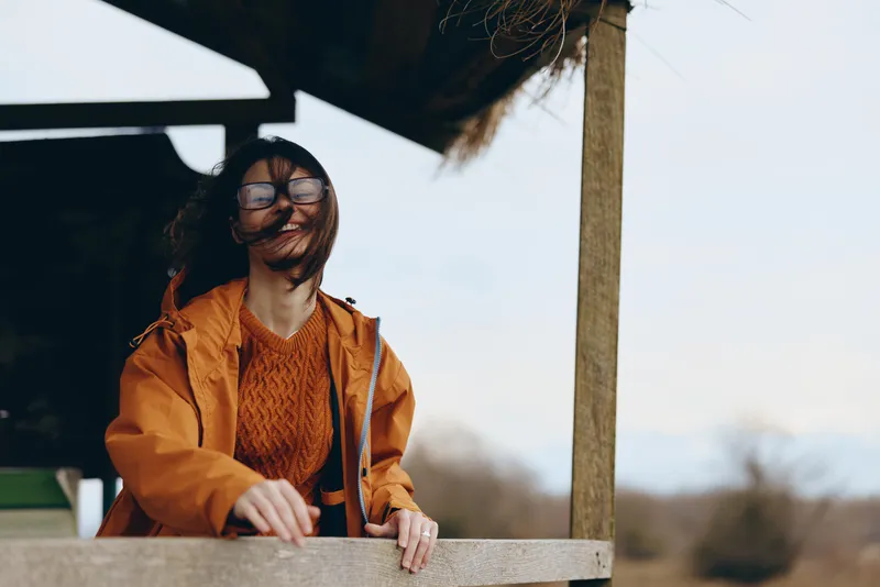 Woman lifestyle portrait outdoors in orange jacket and glasses, smiling with stethoscope and healthcare focus, leaning on wooden railing, sunlight scene with calm countryside background