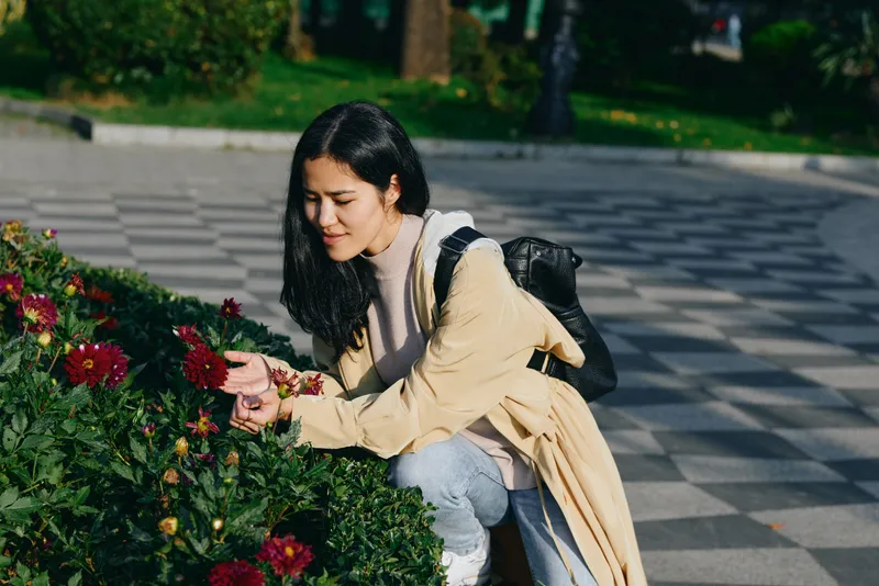 Flowers, woman, park, outdoor, nature, kneeling, contemplation, gardening, petals, bloom, sunlight, the scene captures a peaceful moment as she touches blossoms along a sunny park path