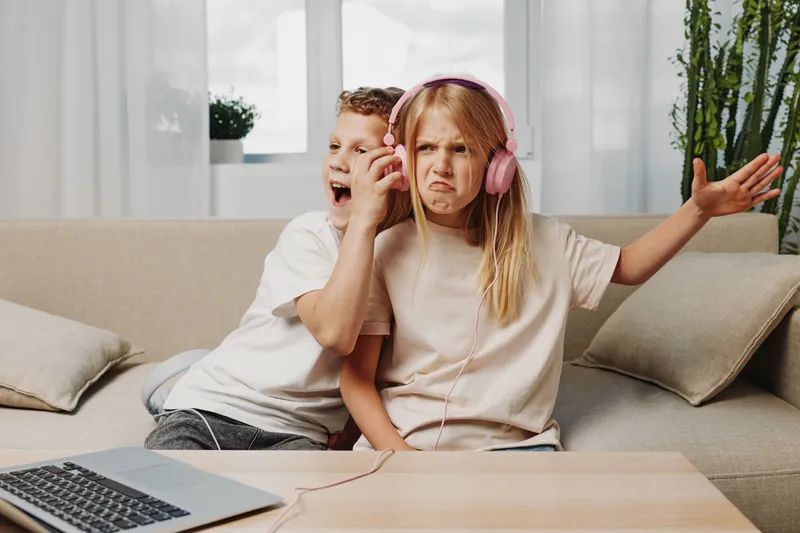 Two children sitting on a couch with one girl wearing pink headphones showing a displeased expression while the boy next to her shouts. Concept of sibling interaction and playful annoyance.