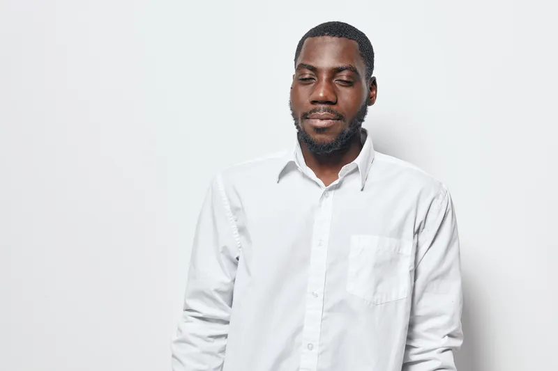 African American man with dark skin wearing white shirt standing with eyes closed against white background. Portrait of calm black male with beard and short hair in casual style.