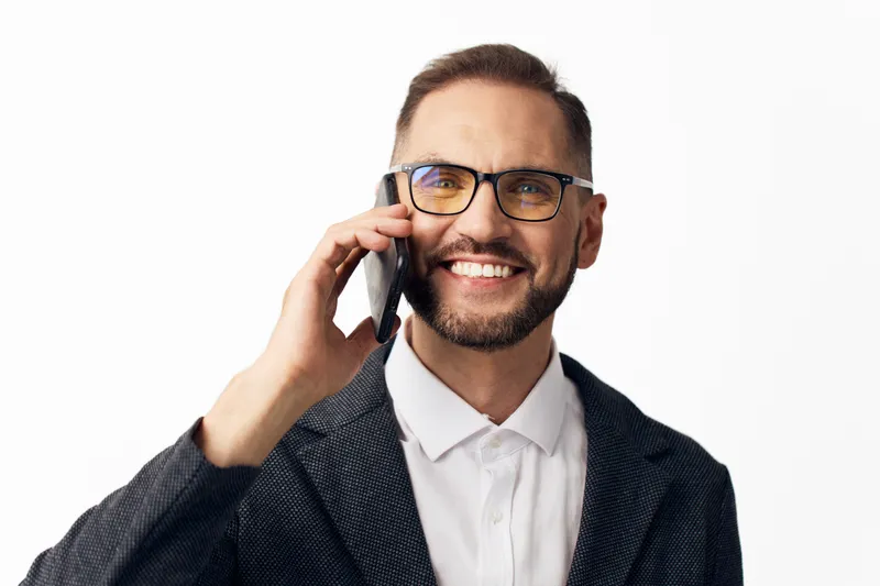 A professional man in business attire smiles while on a cell phone, illustrating confident communication. Isolated colored background highlights a modern, corporate work vibe.