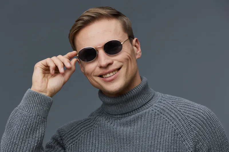 Smiling young man wearing round black sunglasses and a gray turtleneck sweater looks confidently at camera on plain gray background, casual fashion portrait, lifestyle image, autumn or winter season.