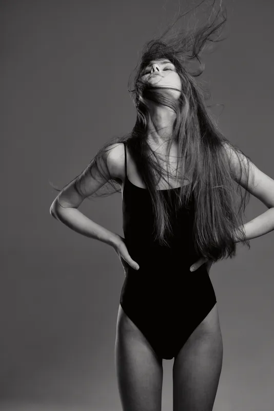 Black and white portrait of a young woman with long hair standing confidently in a black swimsuit with expressive pose and natural movement