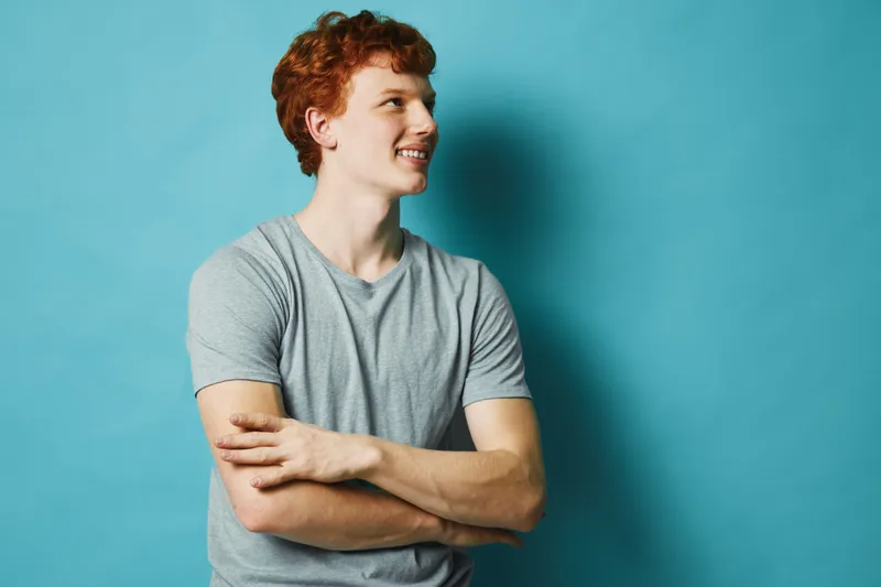 Young man with red hair and freckles wearing gray t shirt looking sideways with crossed arms in front of blue background. Portrait, casual, confident expression in studio lighting.