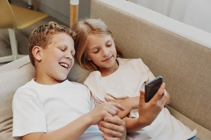 Two happy children sitting on a beige sofa looking at a smartphone together, smiling and enjoying digital entertainment. Concept of friendship, technology, leisure, and childhood fun.