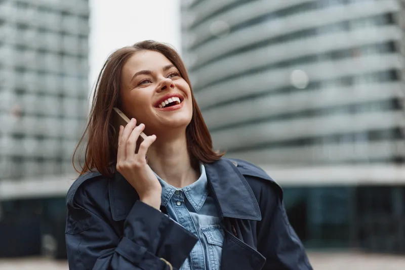 Smiling woman in a denim jacket talks on her mobile on a sunny city street, conveying confidence and a casual, modern vibe.
