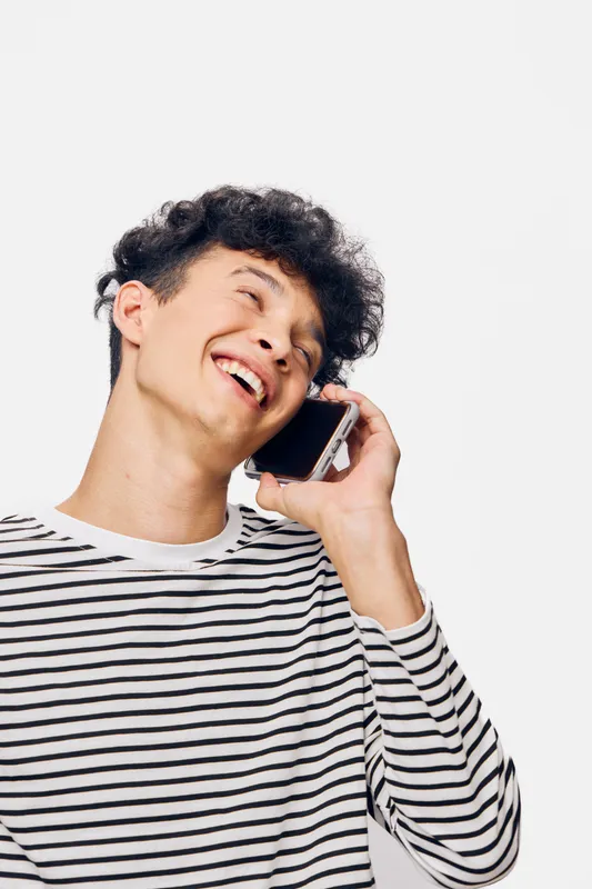 Young man talking on phone, smiling joyfully, wearing striped shirt with curly hair and light skin tone against plain white background, casual lifestyle.