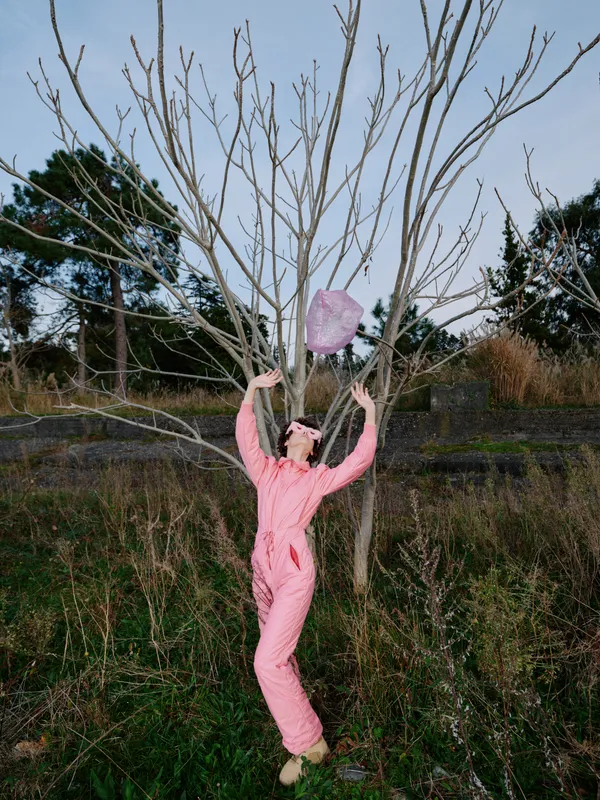 Person in a pink jumpsuit reaches up to a bare tree in an open field, an expressive outdoor moment blending fashion, play, nature, and light among tall grasses