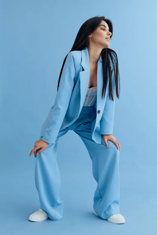 Stylish woman wearing a light blue suit with long straight hair posing confidently against a minimalistic blue background in casual white sneakers.