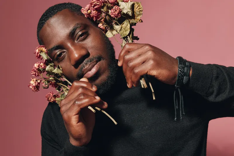 Creative visual designer style portrait of African American man with dark skin holding dried flowers on pink background for artistic expression and fashion concept.