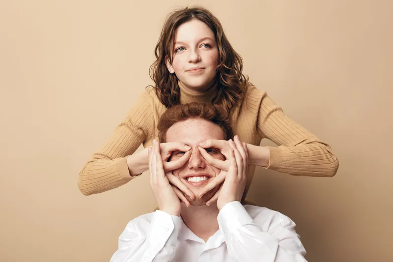 Happy young woman in beige sweater playfully covers smiling man’s eyes with her hands. Studio portrait on beige background. Concept of friendship, fun, and joyful moments together.