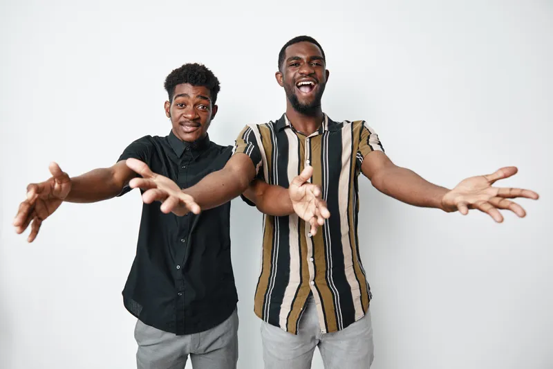 Two African American men with dark skin smiling and reaching out with open arms for hug or welcome on white background. Casual clothing and friendly gesture for connection.