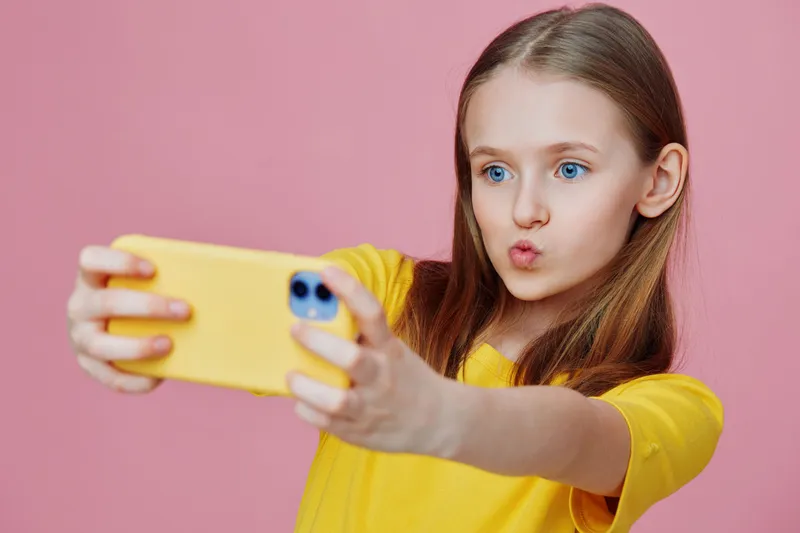 Cute girl taking a selfie with a yellow phone, wearing a bright yellow shirt, with playful expression against a pink background, perfect for social media concepts