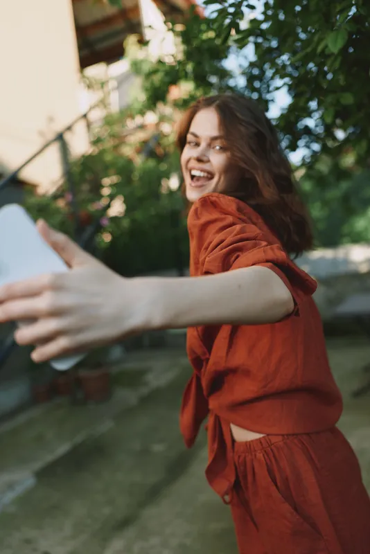 Cheerful woman outdoors, reaching with a paper, red outfit, bright smile, dynamic movement, sunlit scene, natural colors, casual lifestyle in a lush backdrop