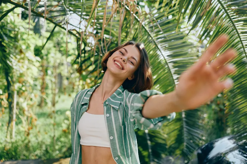Woman, outdoor, smiling, carefree, lush greenery, tropical palm leaves, sunlit scene, casual style, bright smile, confident pose, friendly energy, nature connection, vibrant portrait.