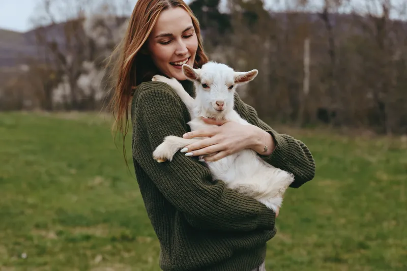Lifestyle scene with a woman in a knitted sweater gently holding a young goat kid in a green field, warm outdoor moment, affectionate gaze, natural light, calm countryside atmosphere