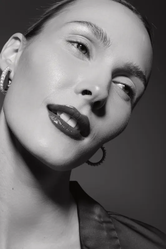 Close-up of a young white woman with clear skin wearing hoop earrings and glossy lipstick, posing in black and white.