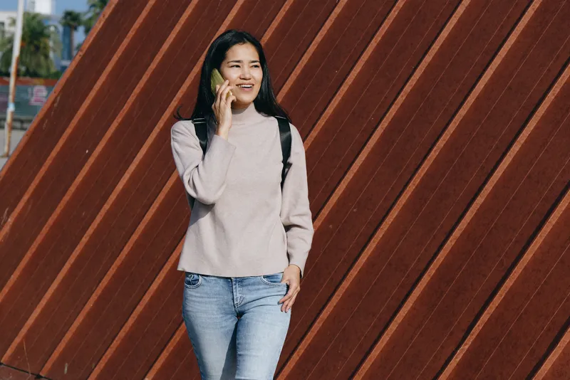 Young woman talking on phone outdoors, casual sweater and jeans, standing beside a diagonal wooden fence, capturing everyday communication and urban street fashion