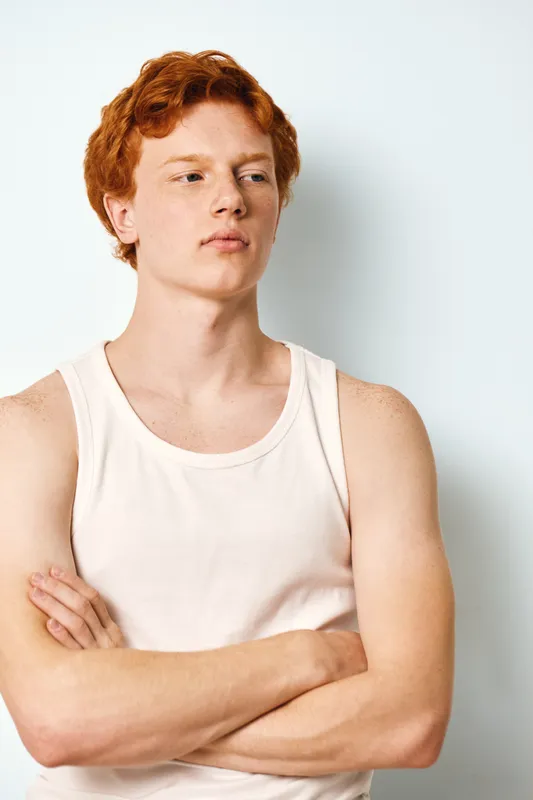 Young man with red hair and freckles, wearing white tank top, standing with arms crossed against white background. Portrait expressing confidence and contemplation.