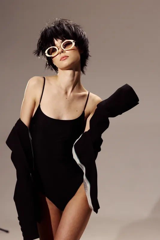 Stylish woman in black bodysuit and sunglasses poses with confident attitude in studio lighting