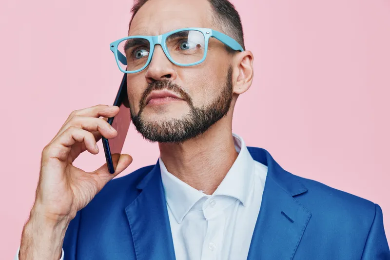 A business professional man standing against a vibrant pink background, speaking on a phone with thoughtful confidence, showcasing focus, determination, and clear emotion.
