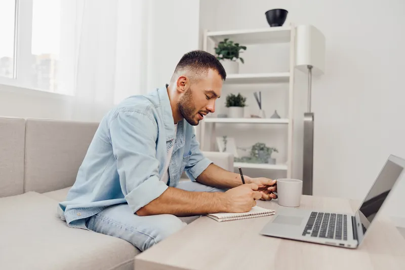 Focused man working at home, jotting down ideas in a notebook, surrounded by a cozy and modern setting. A blend of productivity and comfort is evident in this scene.
