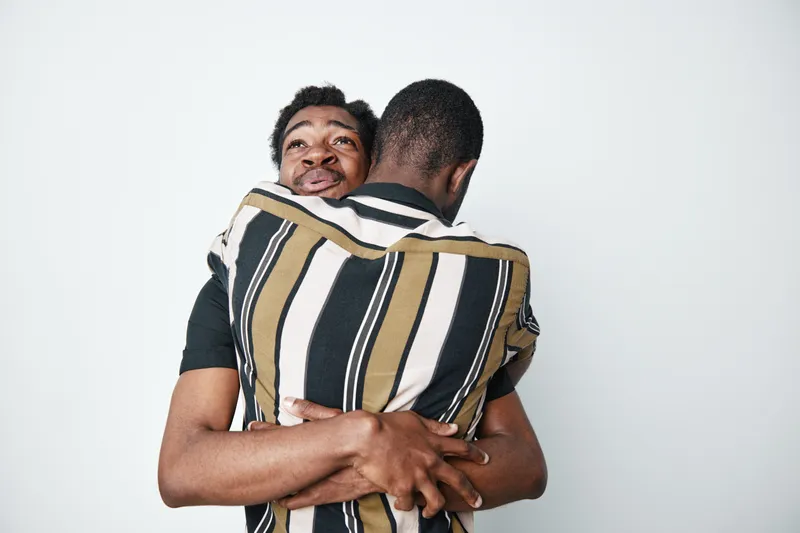 Two young black men with dark skin hugging each other in striped shirts, showing friendship and support in a close embrace against a white background.