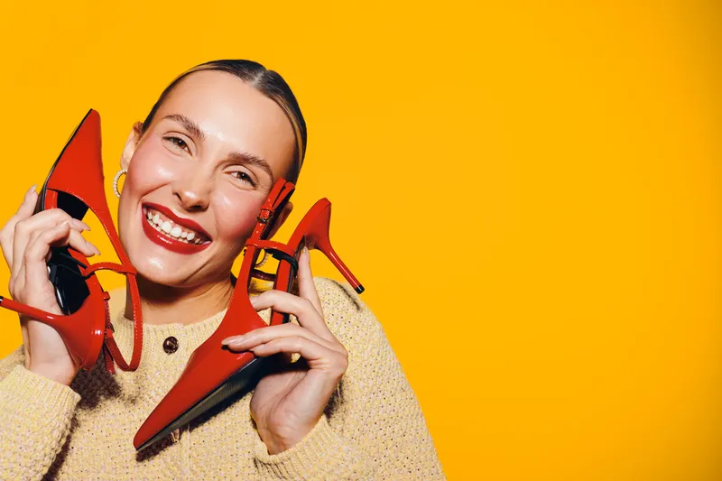 Happy woman with red lipstick holds red high heel shoes near face smiling against bright yellow background. Concept of fashion, style, beauty, and joyful emotions.