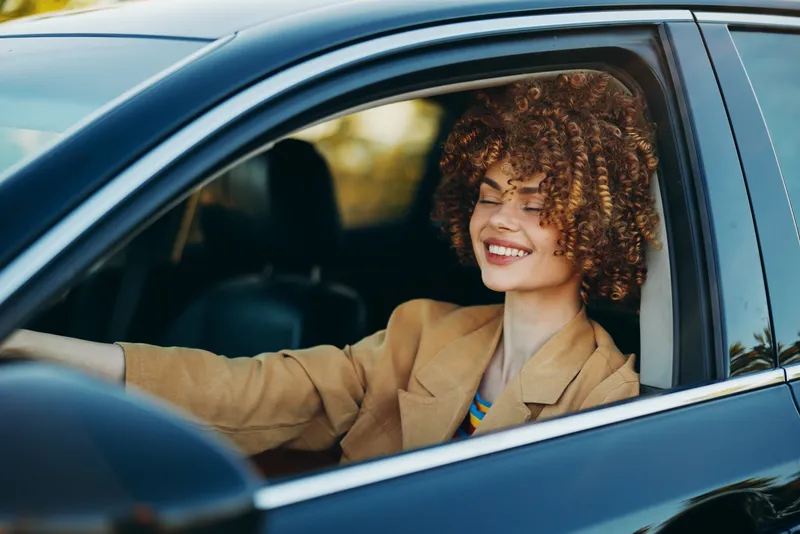 Smiling woman driving a car with curly hair and a tan blazer. Closeup through the window. Concept of travel, style, and daily mobility. Retro film color enhances a warm, nostalgic mood.