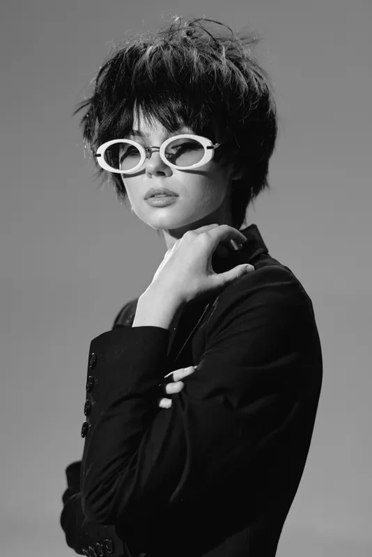 Black and white portrait of a woman wearing white sunglasses and a stylish blazer with a short hairstyle and confident expression.