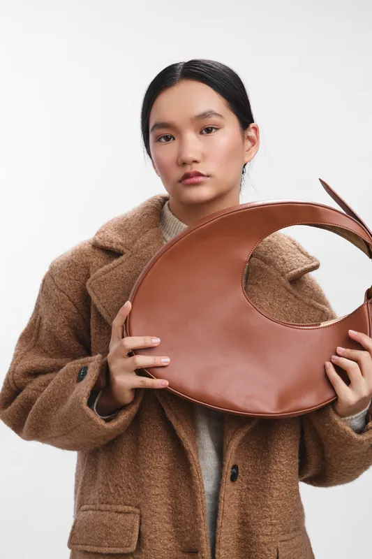 asian woman stands in a neutral studio holding a large circular leather bag handle, presenting a fashion accessory with a calm, poised expression.