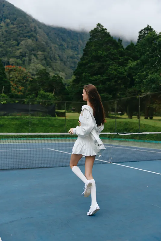 A young woman in a stylish white tennis outfit runs gracefully on a tennis court surrounded by lush greenery, embodying energy and confidence