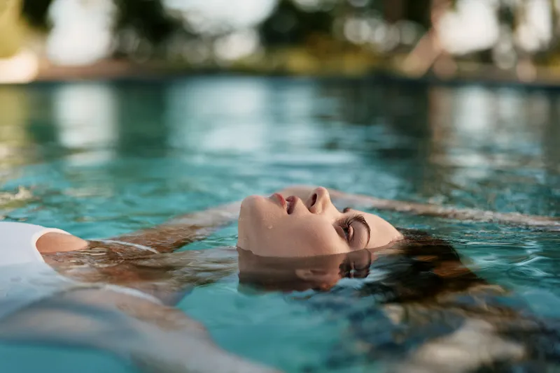 A woman floats in a sunlit pool, eyes closed, conveying relaxation, calm, and leisure in an outdoor water scene that captures tranquil lifestyle and restorative wellness.