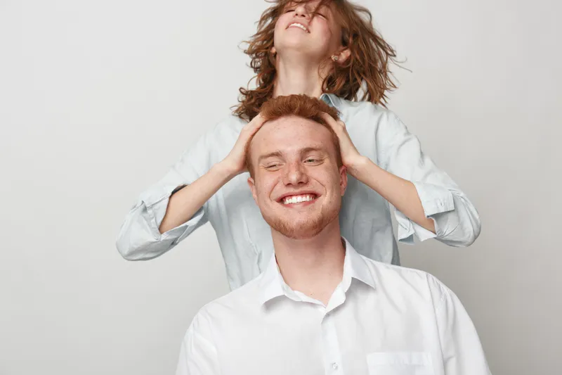 Happy young man with red hair smiles while woman with curly hair playfully covers his eyes. Light background, casual clothing, concept of friendship and joyful moments together.