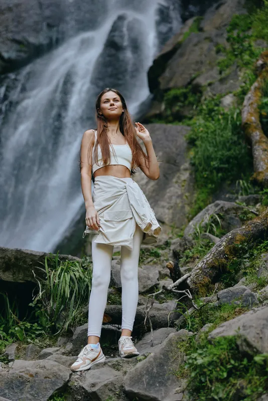 young woman standing near waterfall in activewear with hiking shoes, surrounded by green nature and rocks, outdoor adventure and fitness concept