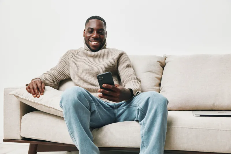 Happy African American man in cozy sweater using smartphone at home, relaxed atmosphere and casual style, enjoying leisure time on sofa