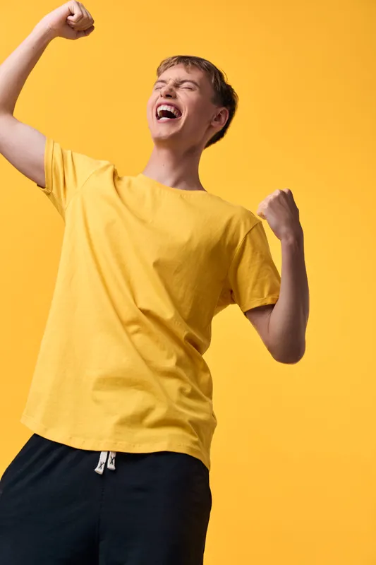 Celebration mood with energetic man in yellow shirt, fists raised and mouth open in joy, bright studio backdrop signaling victory and momentum for success