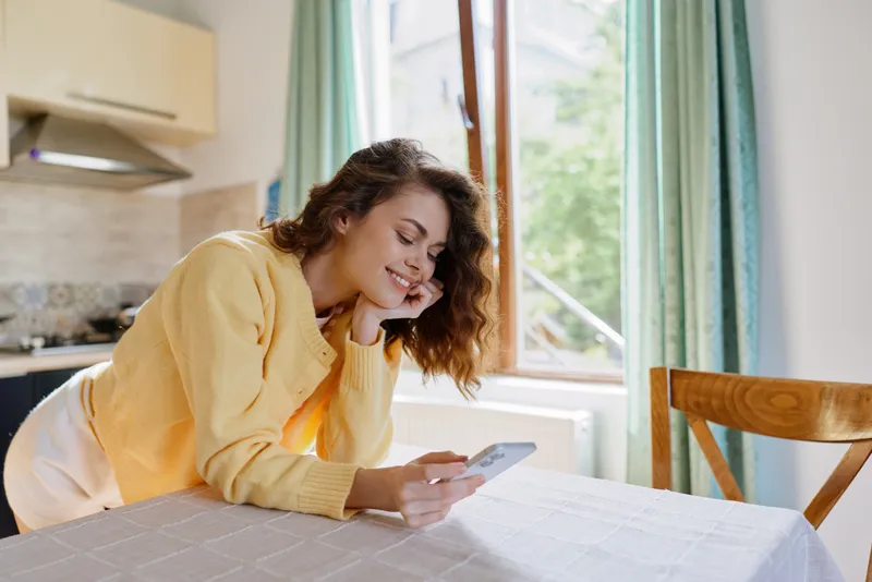 Cheerful woman at home, relaxing with smartphone, daylight through window, casual sweater, cozy kitchen, lifestyle moment, natural colors, candid indoor scene, relaxed posture