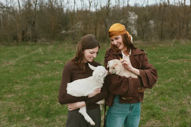 Two women, friends, outdoors in a grassy field cuddle goat kids, sharing smiles. A lifestyle moment with animals and rural scenery, highlighting care, connection, and gentle play in a countryside
