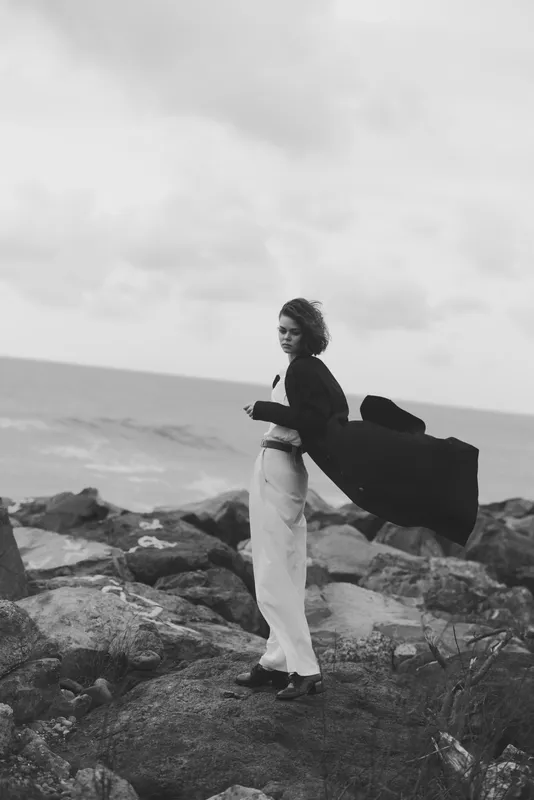 Woman in a cape stands on rocky coast, fashion forward pose in monochrome setting with wind lifting fabric, rugged background highlights style and confidence.