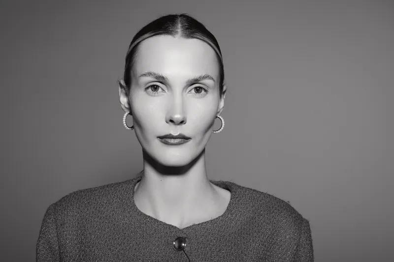 Portrait of a serious white woman in her thirties with light skin and slicked back hair wearing a textured jacket against a plain background.