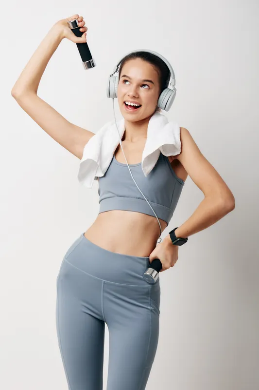 Fitness-conscious woman in gray athletic wear lifts a dumbbell with one arm, headphones on and a towel over her shoulder, posing in a clean studio to convey strength and wellness during a workout.