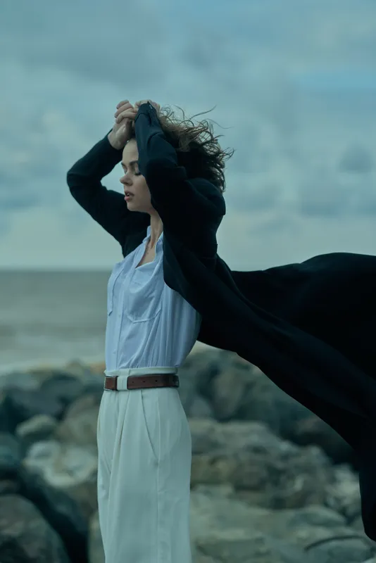Wind, coat, flowing, sea breeze, model on a rocky coastline. Fashionable garment in motion against a moody seascape and cloudy sky, showcasing dynamic elegance and freedom.