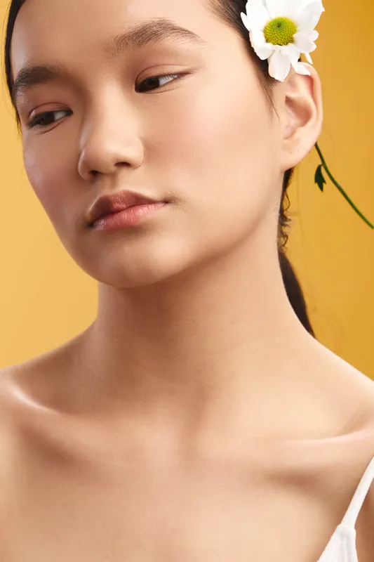 Asian woman gazes softly to the side against a warm, neutral backdrop, showcasing clear skin and subtle makeup in a refined portrait that conveys quiet elegance and poise.