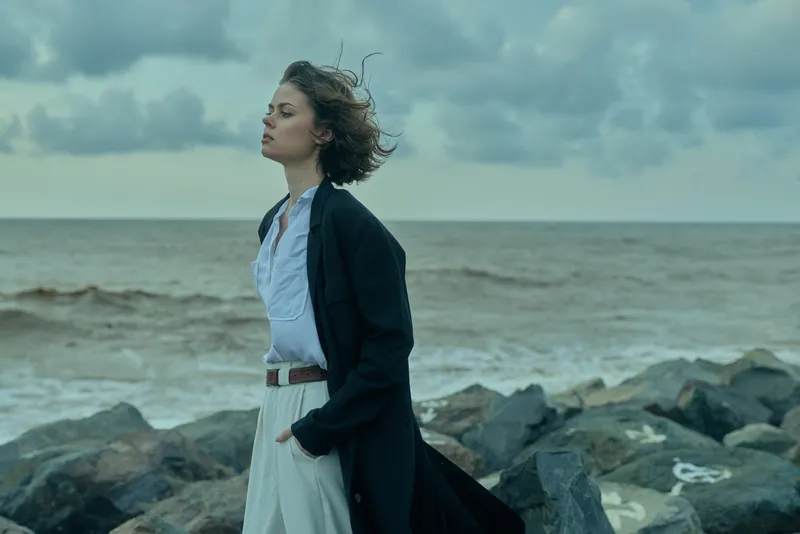 Woman on rocky coast with windblown hair, wearing a long coat, by the sea under a moody sky, conveying fashion, outdoor mood and dramatic coastal style
