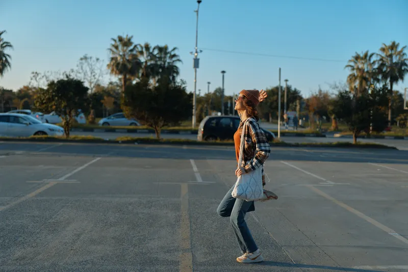 Woman walking candid through an urban parking lot at golden hour with palm trees and casual outfit, backpack and movement capture authenticity, mindful living and emotional storytelling.