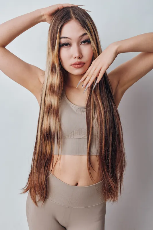 Young woman with long straight hair posing in neutral colored workout outfit against light background. Casual style, fitness clothing, confident look and natural makeup.